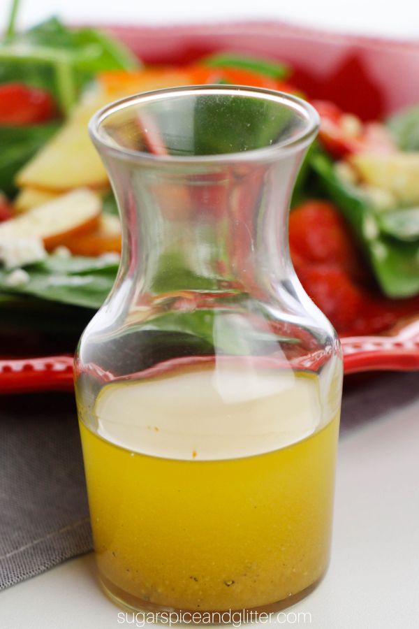 A small karafe of apple cider vinagrette dressing in front of a red plate with salad