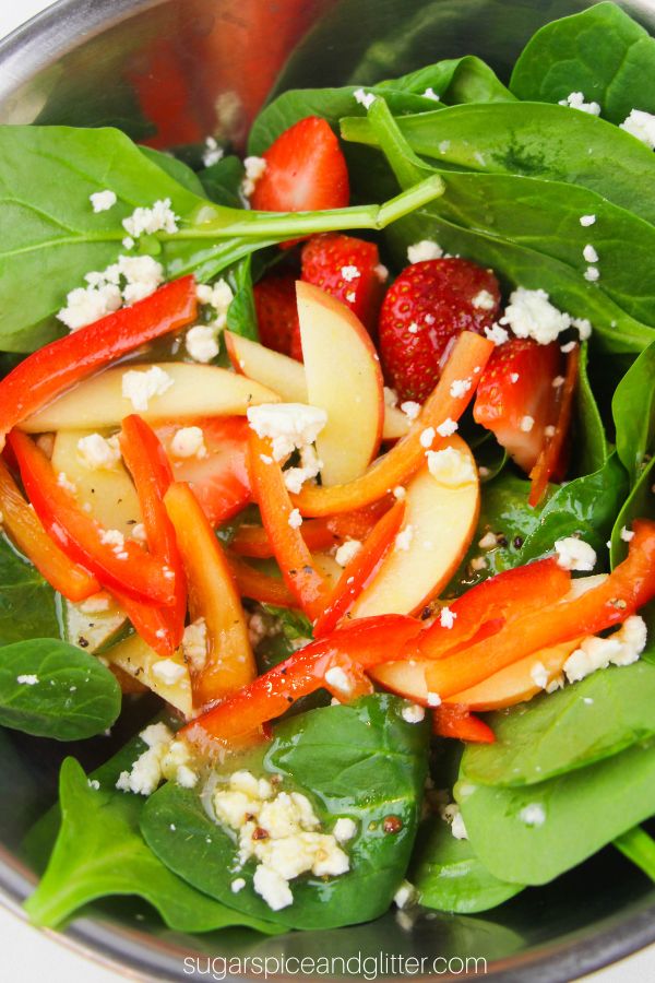 A close-up picture of an apple, roasted red pepper, strawberry and feta salad with spinach drizzled with apple cider vinaigrette