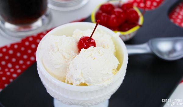 overhead picture of root beer ice cream in a white pedestal bowl with a cherry on top and mugs of root beer and a bowl of cherries in the background
