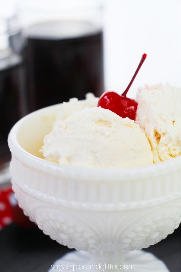 a close-up picture of root beer ice cream in a white pedestal bowl with a cherry on top and mugs of root beer in the background