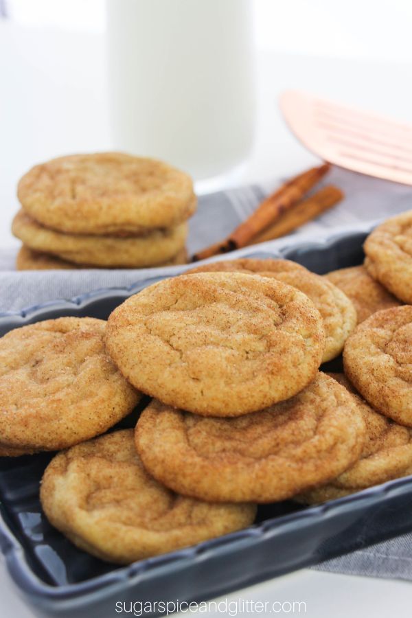 a blue tray piled high with golden brown snickerdoodle cookies, with a glass of milk, cinnamon sticks and a stack of more cookies in the background