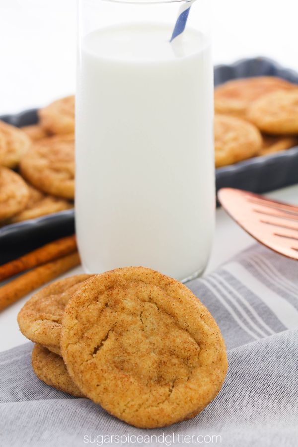 a stack of golden snickerdoodle cookies with a glass of milk and a tray piled high with even more snickerdoodle cookies in the background