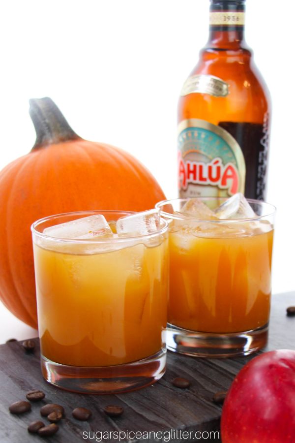 Two glasses of apple cider cocktails on a gray slate tray, with coffee beans scattered around, a red apple in the forefront and a bottle of Kahlua and a pumpkin in the background