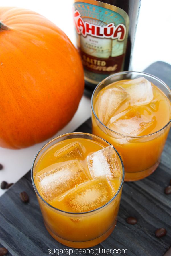 Two glasses of apple cider cocktails on a gray slate tray, with coffee beans scattered around, a bottle of Kahlua and a pumpkin in the background