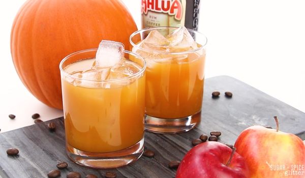 Two glasses of apple cider cocktails on a gray slate tray, with coffee beans scattered around, a red apple in the forefront and a bottle of Kahlua and a pumpkin in the background