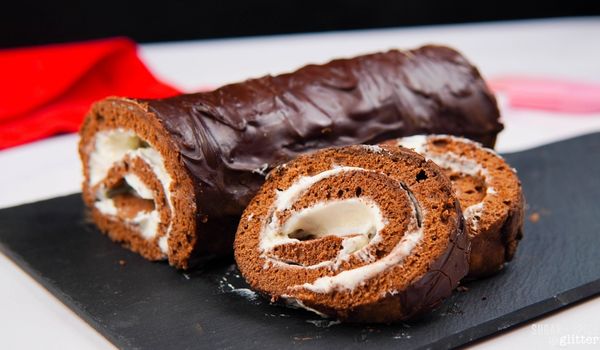 Picture of chocolate roll cake with two slices placed in front of the roll, on top of a black cutting board