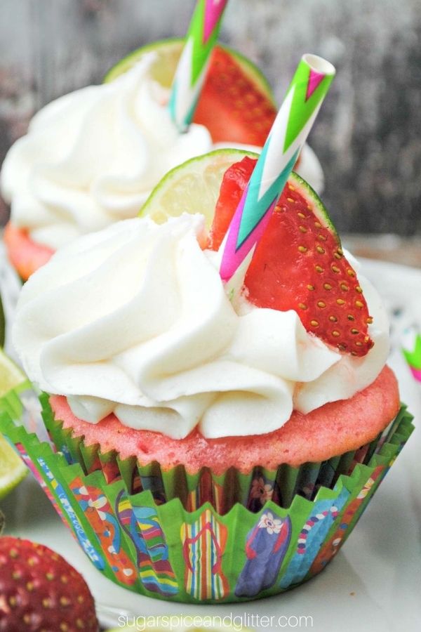 a white platter with strawberry cupcakes with white frosting garnished with a slice of strawberry and a slice of lime and a small straw stuck into the frosting