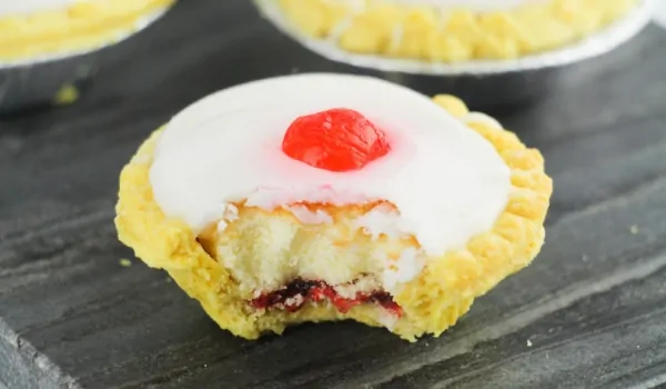 Cherry bakewell tarts on a slate tray with a bite out of it showing all of the different layers - pie crust, jam, almond cake and frosting