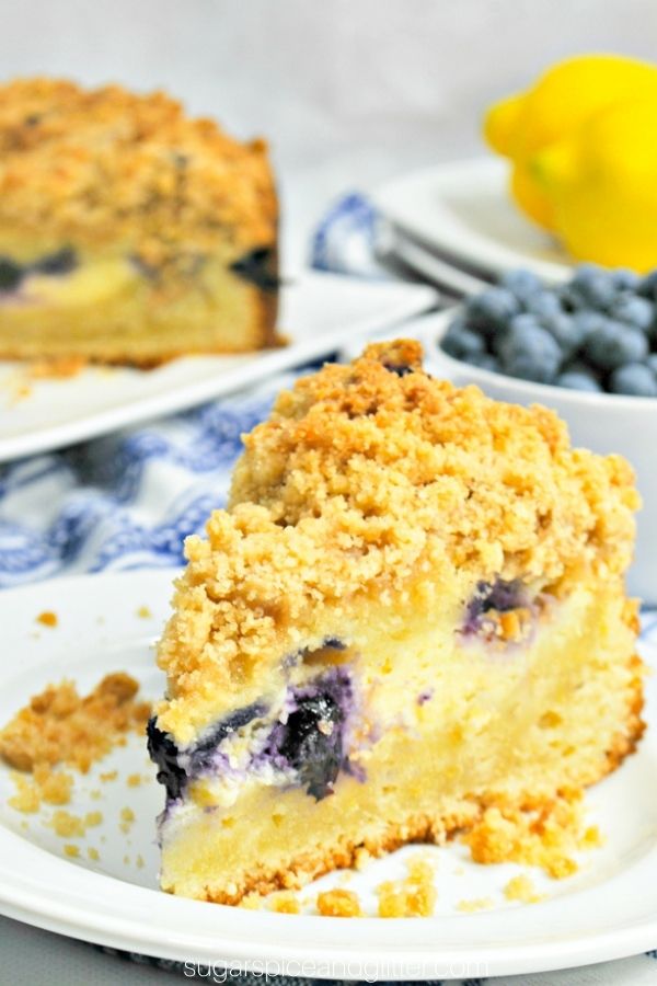 a slice of lemon blueberry coffee cake on a white plate with a bowl of blueberries and the whole cake in the background