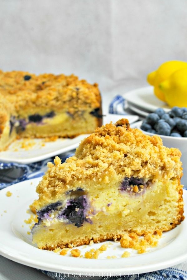 a slice of lemon blueberry coffee cake on a white plate with a bowl of blueberries and fresh lemons in the background