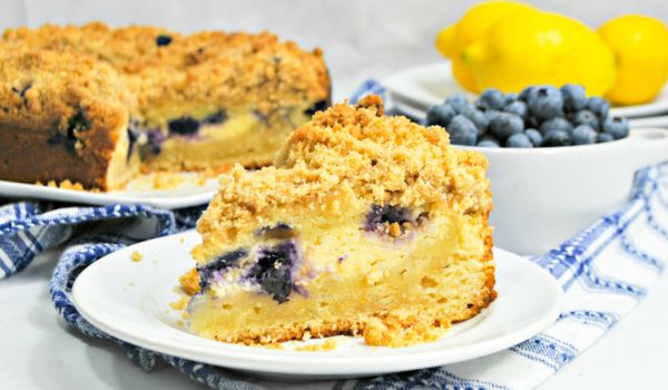 a slice of lemon blueberry coffee cake on a white plate with a bowl of blueberries and fresh lemons in the background