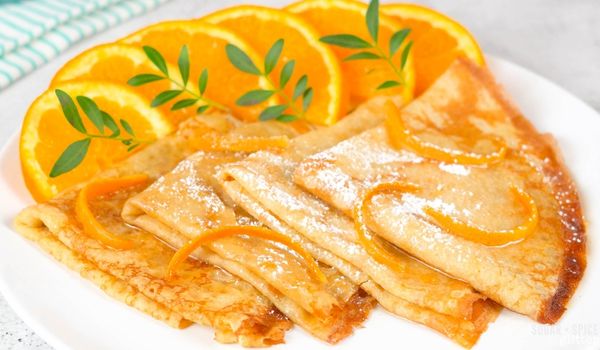 white plate of folded crepes soaked in orange sauce sprinkled with orange peels and powdered icing sugar