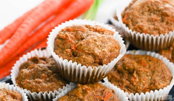 Close-up of a carrot muffin in a white muffin liner on top of a pile of carrot muffins with a bunch of carrots in the background