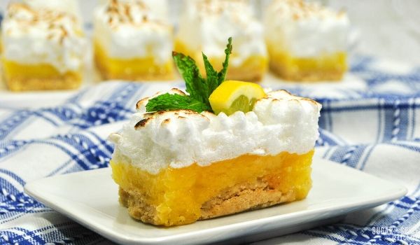 a slice of lemon meringue pie bar with a slice of lemon and fresh mint on top placed on a white plate with more lemon pie bar slices in the background
