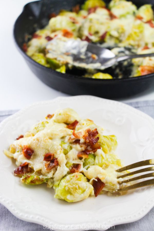Cheese and bacon covered brussels sprouts on a white plate set against a gray napkin with a cast-iron skillet with more brussels sprouts in the background