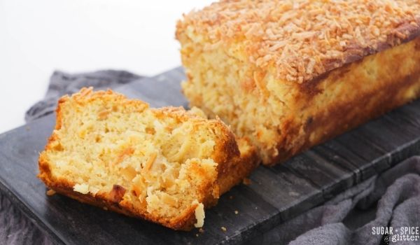 a loaf of pineapple coconut bread on a gray slate tray with two slices cut out and displayed to show the pineapple and coconut speckled throughout the loaf