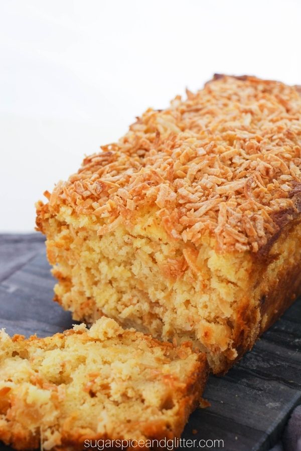 a loaf of pineapple coconut bread on a gray slate tray with two slices cut out and displayed to show the pineapple and coconut speckled throughout the loaf