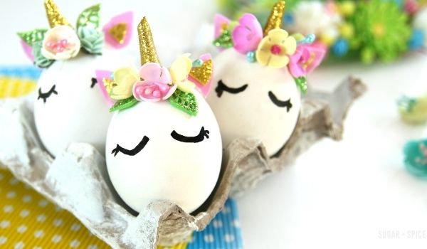three Unicorn Easter Eggs decorated with a unicorn horn, ears and a flower crown set inside an egg carton with flowers scattered around