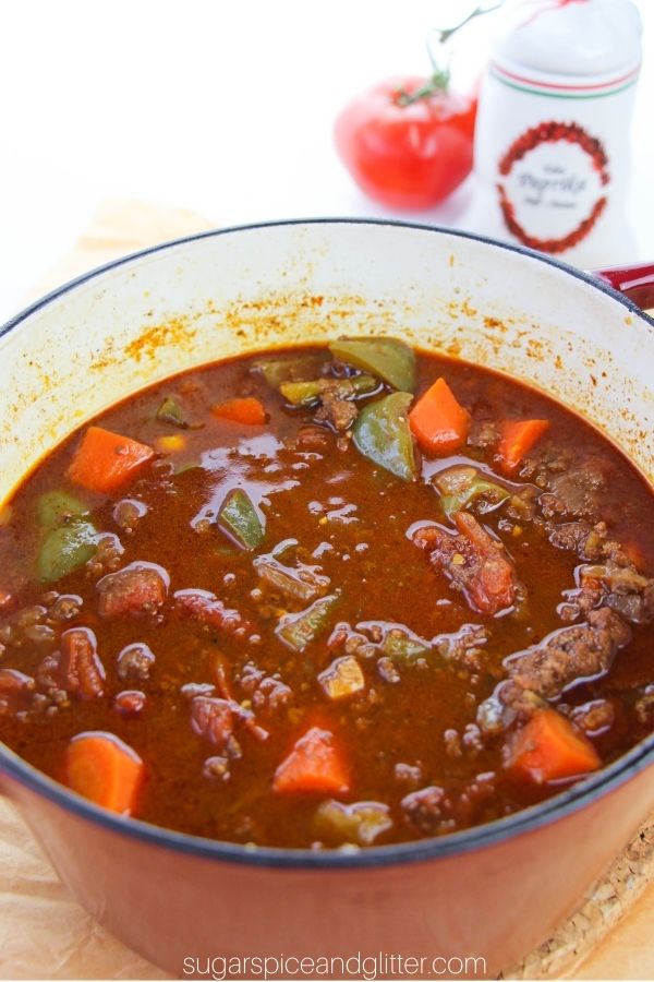close-up shot of a red dutch oven filled with beef and vegetable stew with paprika and tomatoes in the background