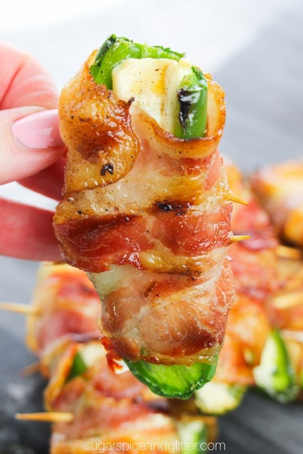 hand holding up a bacon-wrapped jalapeño popper over a tray holding more bacon-wrapped jalapeño poppers