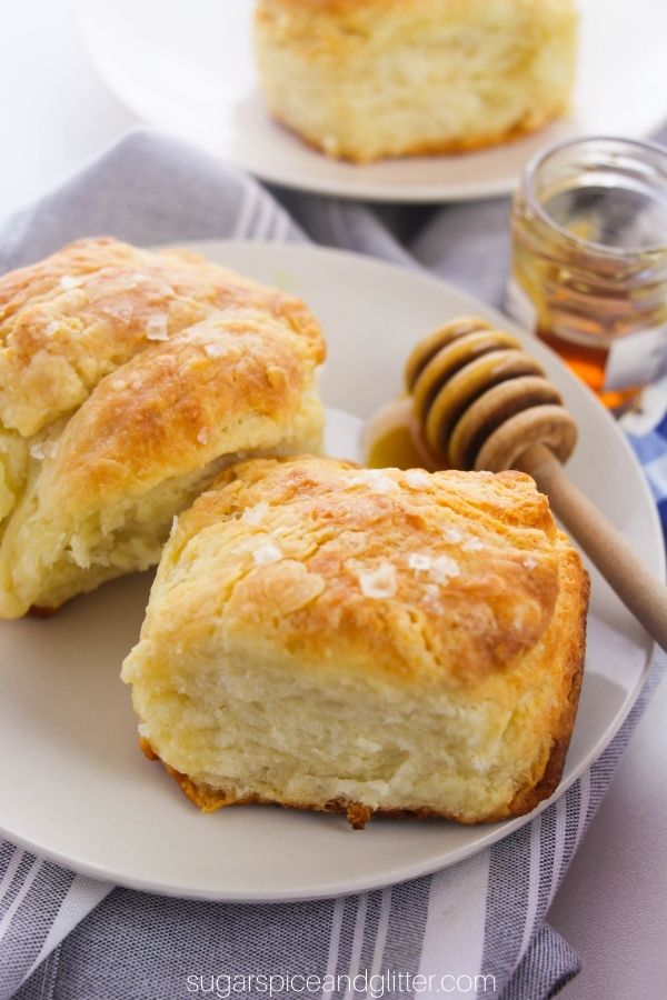 golden brown buttermilk biscuits with flaked salt on top and a honey dipper set to the side on a white plate, with a jar of honey in the background
