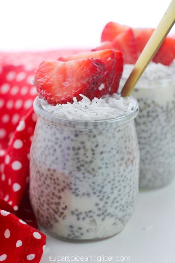 two glass jars filled with chia seed pudding, topped with shredded coconut and sliced strawberries with a red polka dot napkin in the background