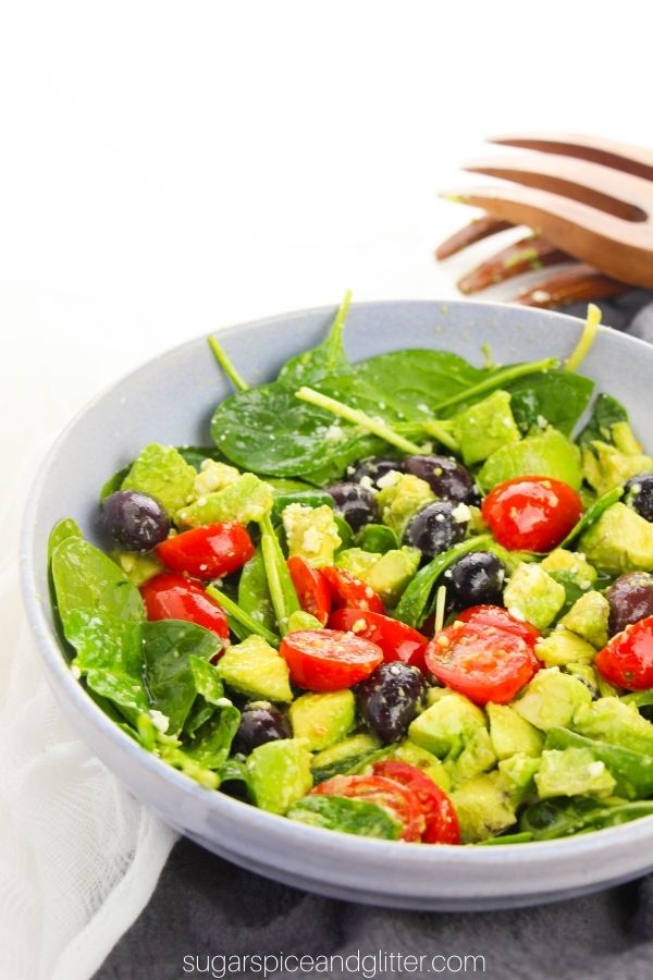 a large blue bowl full of Greek spinach salad on a gray napkin with wooden salad hands in the background