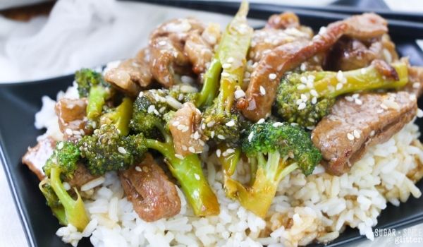 beef and broccoli stir fry on a bed of white rice on a square, black plate with chopsticks on the edge on a white napkin