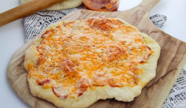 baked cheese pizza on a wooden pizza peel with ingredients in the background