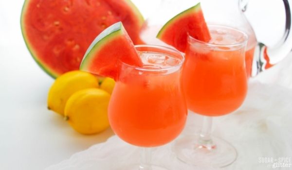 two mini-hurricane glasses filled with ice and watermelon lemonade garnished with wedges of watermelon, with a cut watermelon, lemons and a pitcher of lemonade in the background