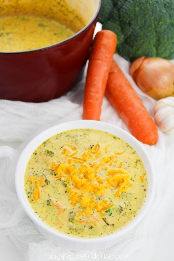 overhead image of a white bowl of broccoli cheddar soup along with a dutch oven filled with more soup and the ingredients needed to make the soup in the background