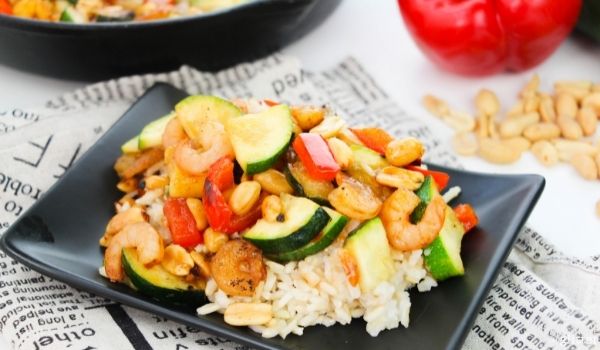 a black square plate with rice topped with Kung Pao Shrimp, with ingredients needed to make the recipe in the background
