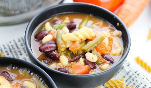two black bowls filled with minestrone soup with pasta noodles scattered around