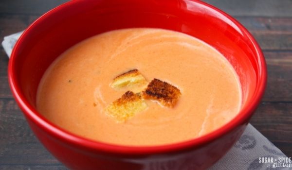 a red bowl filled with creamy tomato soup with a few grilled cheese croutons sprinkled on top