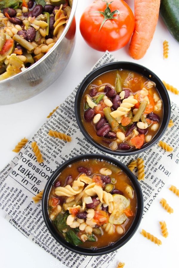 overhead image of two black bowls filled with minestrone soup with pasta noodles scattered around