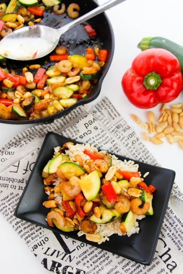 a black square plate with rice topped with Kung Pao Shrimp, with a skillet holding more of the stir-fry and the ingredients needed to make the recipe in the background