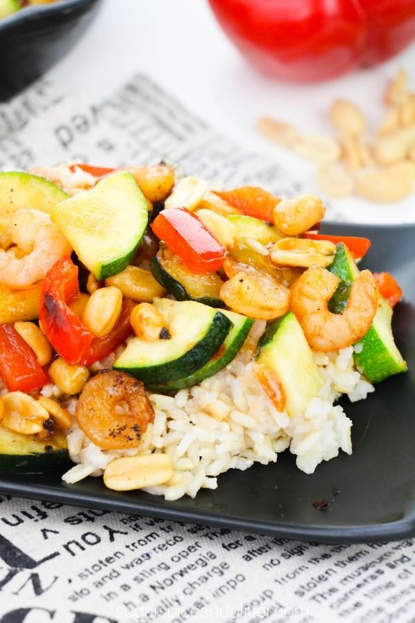 a black square plate with rice topped with Kung Pao Shrimp on a newsprint napkin