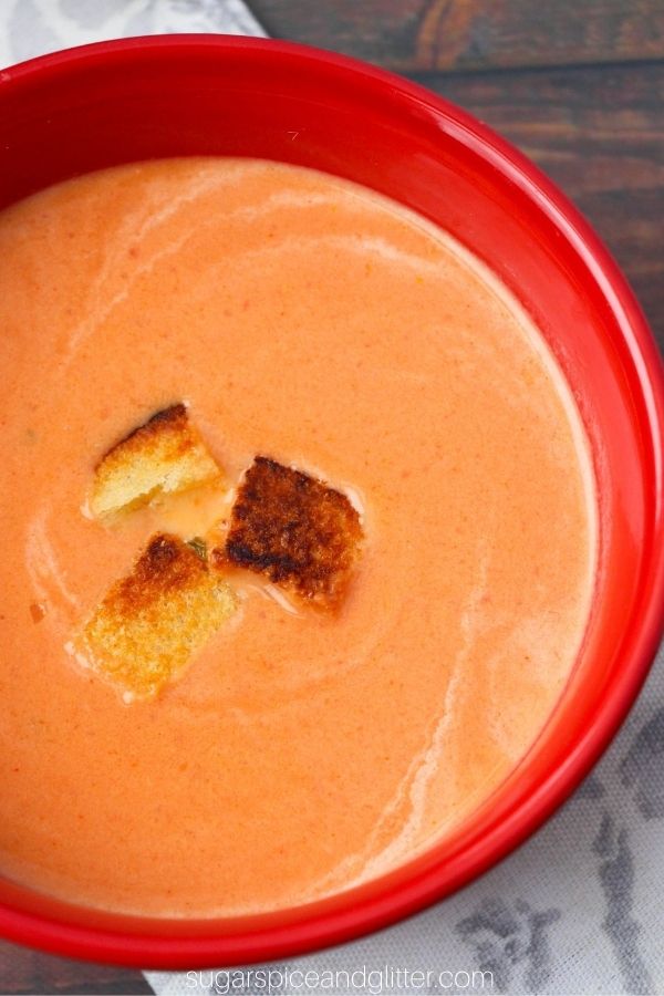 overhead close-up picture of a red bowl filled with creamy tomato soup with a few grilled cheese croutons sprinkled on top