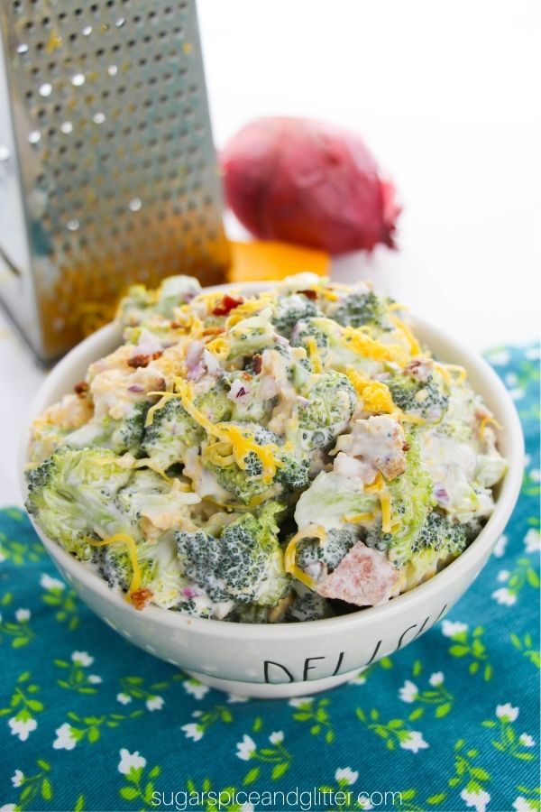 a white bowl filled with broccoli salad set on a blue floral napkin, with cheese and red onion in the background