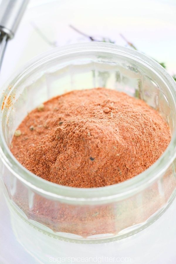 close-up image of a jar holding red blackening spice with a whisk and fresh herbs in the background