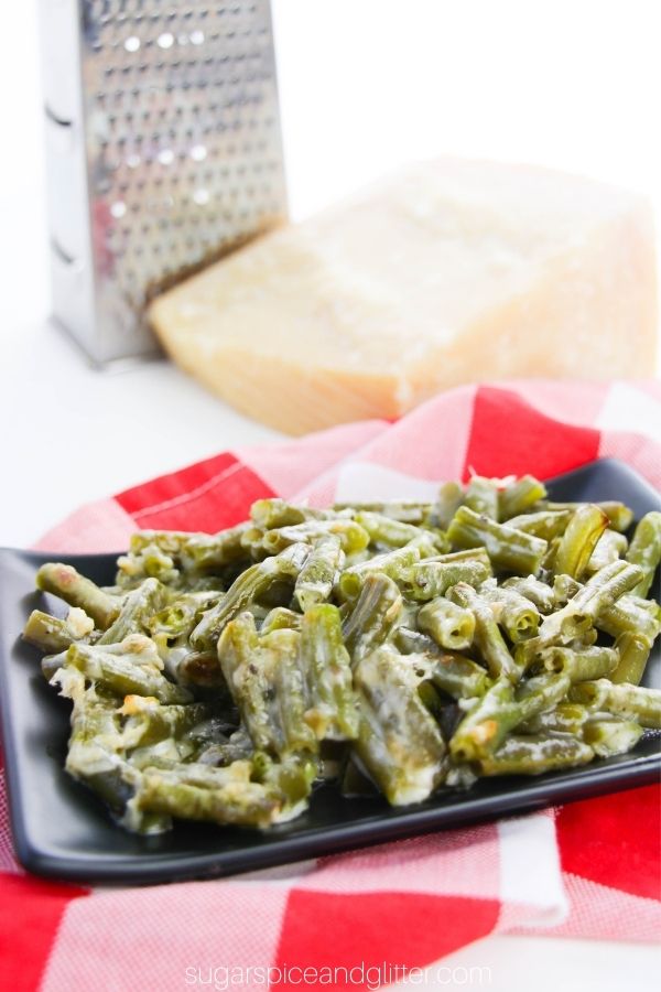 cheesy green beans on a square, black plate set on a red plaid napkin with Parmesan cheese and a grater in the background