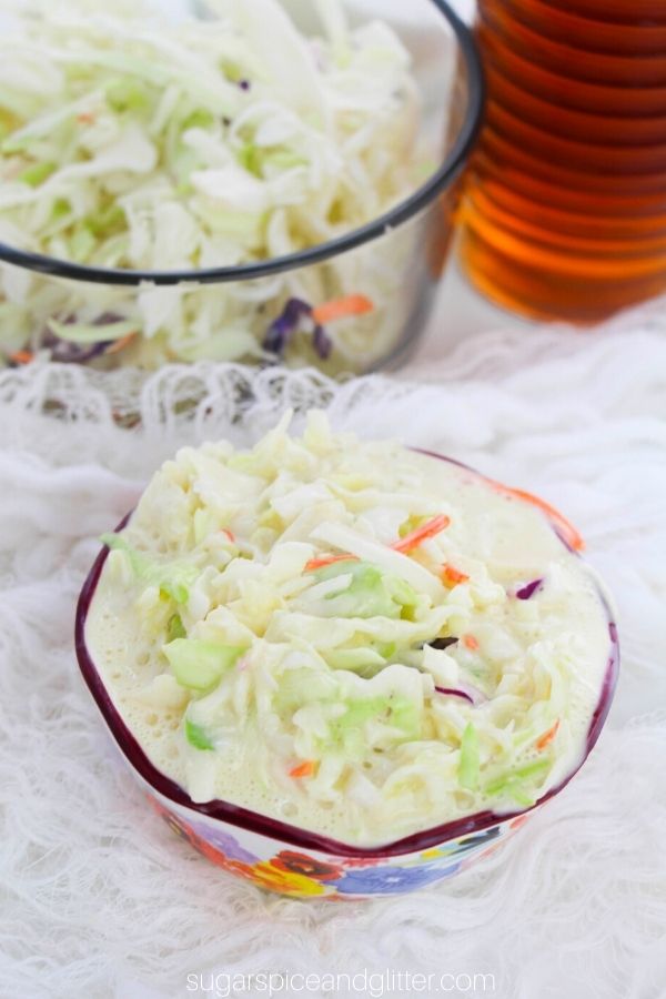 a floral bowl filled with coleslaw on a white muslin napkin with a giant mixing bowl of coleslaw and a bottle of apple cider vinegar in the background