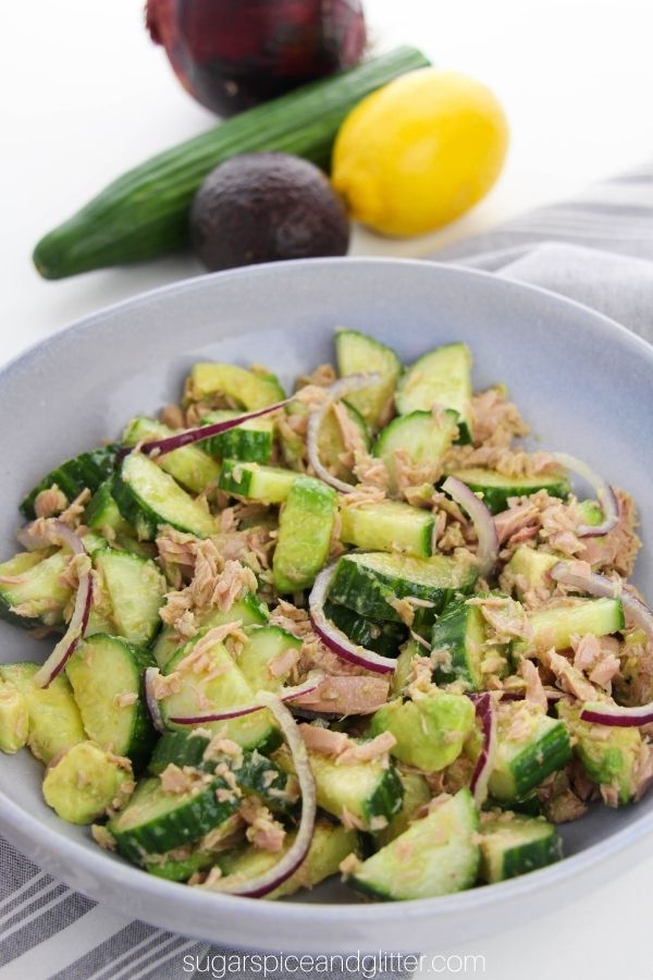 large blue bowl full of cucumber tuna salad with a red onion, cucumber, lemon and avocado in the background