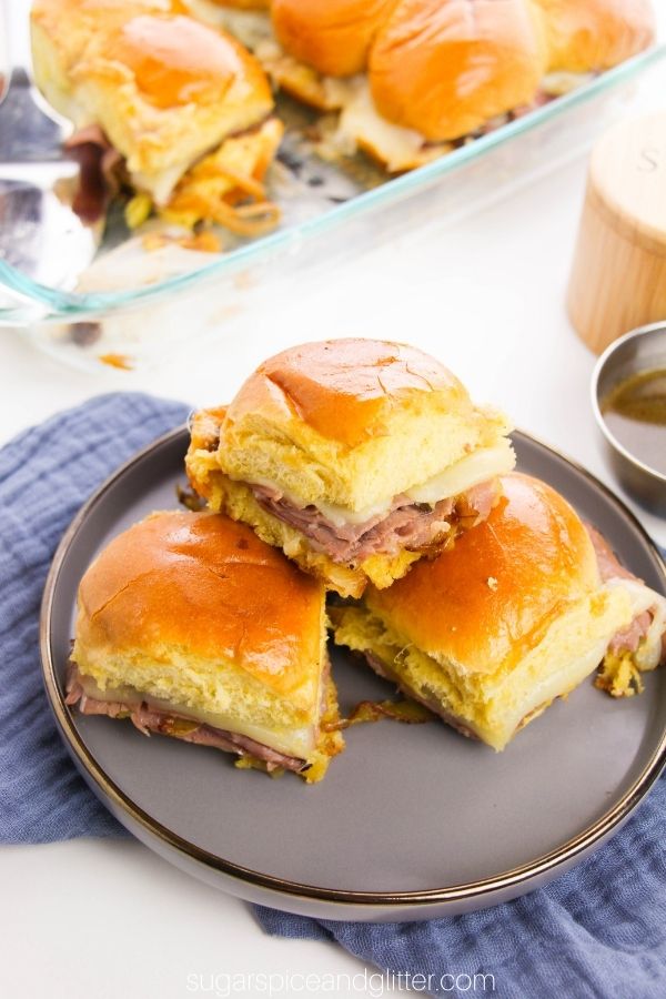 overhead shots of French dip sliders on a gray plate with a silver bowl of au jus dipping sauce to the side and a casserole dish full of sliders in the background