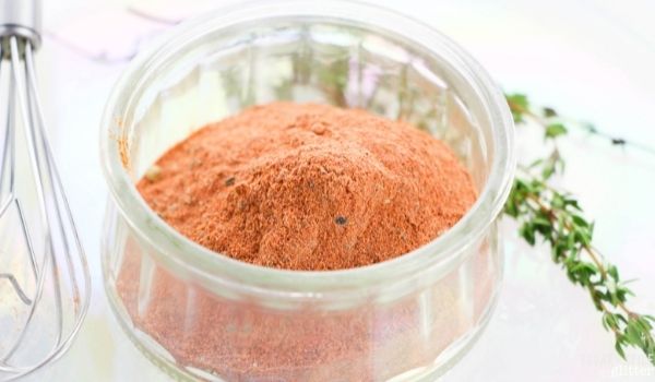 close-up image of a jar holding red blackening spice with a whisk and fresh herbs in the background
