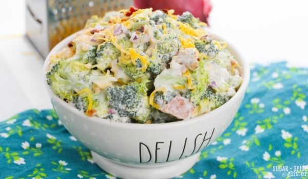 a white bowl filled with broccoli salad set on a blue floral napkin, with cheese and red onion in the background