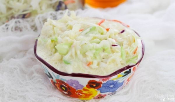 a floral bowl filled with coleslaw on a white muslin napkin with a giant mixing bowl of coleslaw and a bottle of apple cider vinegar in the background