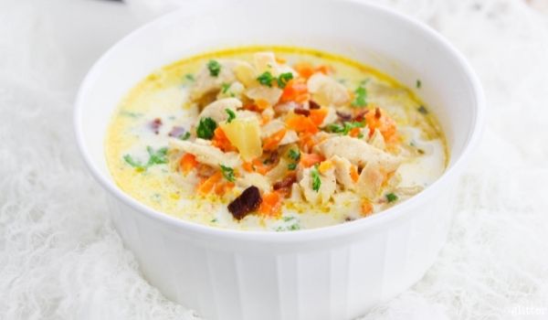 a white bowl full of cheddar bacon ranch chicken soup with ingredients needed to make the soup in the background plus a large dutch oven full of more soup