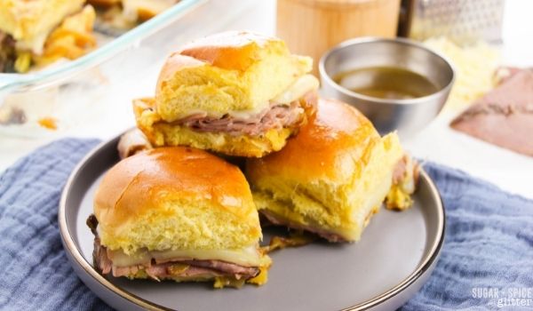 French dip sliders on a gray plate with a silver bowl of au jus dipping sauce to the side and a casserole dish full of sliders in the background along with ingredients used to make the sliders