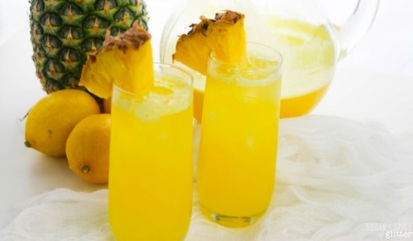 two glasses of pineapple lemonade garnished with pineapple wedges, with a pineapple, lemons and a pitcher of the lemonade in the background